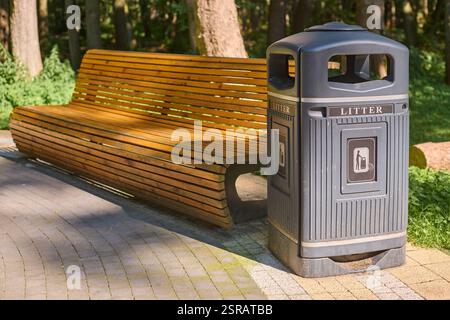 Parkbank aus Holz neben Abfalleimer auf sonnendurchflutetem Steinweg in ruhiger Waldlage, einladende Erholung und achtsame Entsorgung. Friedliches Ambiente in üppiger Umgebung Stockfoto