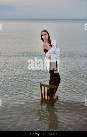Die junge Frau steht selbstbewusst auf einem Stuhl, getaucht in ruhiges, flaches Meer. In lässiger Kleidung strahlt sie Ruhe aus vor dem weiten Horizont. Stockfoto