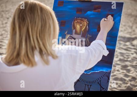 Junge Künstlerin, die mystische Porträts auf Leinwand am ruhigen Strand malte. Das Kunstwerk zeigt ein surreales weibliches Gesicht, umgeben von abstrakten Elementen, die evozieren Stockfoto