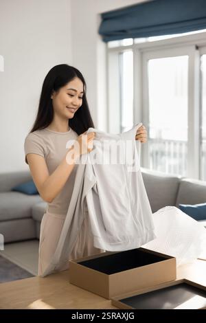 Die junge asiatische Frau, die beim Auspacken des Paketbogens mit Kleidung ankommt Stockfoto
