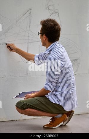 Junger kaukasischer Mann mit Brille hockt sich, skizziert an einer großen Außenwand. Gestreiftes Hemd und grüne Shorts vor hellem, neutralem Hintergrund. Fokussiert Stockfoto