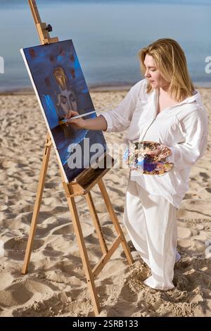 Junge Künstlerin, die mystische Porträts auf Leinwand am ruhigen Strand malte. Das Kunstwerk zeigt ein surreales weibliches Gesicht, umgeben von abstrakten Elementen, die evozieren Stockfoto