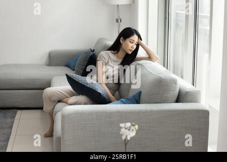 Nachdenkliche traurige asiatische Frau, die im Wohnzimmer auf dem Sofa sitzt Stockfoto