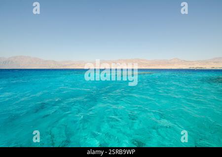 Kristallklares türkisfarbenes Wasser erstreckt sich über das Rote Meer mit entfernten, trockenen Bergen unter wolkenlosem Himmel. Die ruhige Meereslandschaft bietet einen lebhaften Kontrast von se Stockfoto