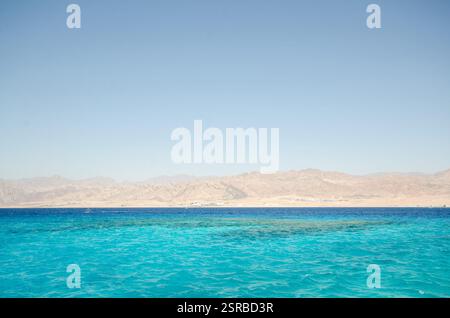 Kristallklares türkisfarbenes Wasser erstreckt sich über das Rote Meer mit entfernten, trockenen Bergen unter wolkenlosem Himmel. Die ruhige Meereslandschaft bietet einen lebhaften Kontrast von se Stockfoto