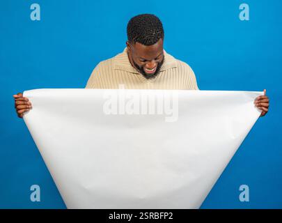 Studio-Aufnahme eines lächelnden Mannes, der ein großes weißes Blatt Papier vor blauem Hintergrund hält Stockfoto