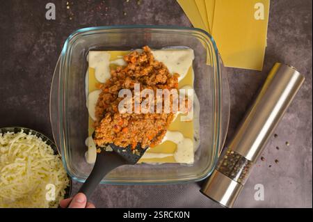 Hausgemachte Lasagne-Zubereitung. Die Hand der Frau verwendet einen Spachtel, um die Bolognese-Sauce in Glasform zu verteilen. Nudelblätter, Béchamelsauce Stockfoto