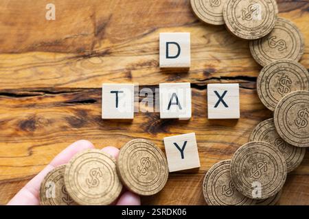 Hölzerne Buchstaben mit der Aufschrift „STEUERTAG“ auf einer strukturierten Holzoberfläche, umgeben von Münzen mit Dollarzeichen. Steuern, Finanzen und wirtschaftliche Verantwortung Stockfoto