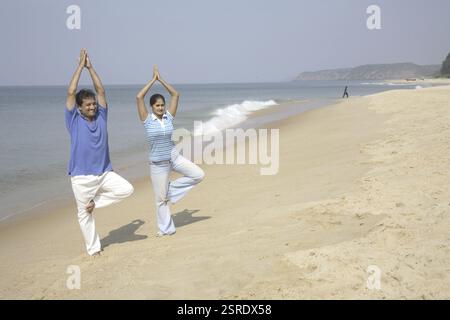Südasiatische indische junge Mann und Frau, die Yoga machen, stehen auf einem gefalteten Bein und heben Gelenkhandhandflächen über der Küste, Shiroda, Dist. Sindhu Stockfoto