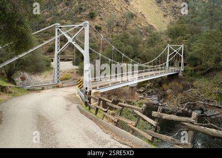 Die alte Hängebrücke überspannt einen Fluss. Straßenkurven darunter, abgenutzter Holzzaun daneben. Briceburg Suspension Bridge, Mariposa County, Kalifornien Stockfoto