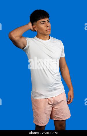 Junger latino-Mann mit weißem T-Shirt und rosa Shorts, der sein Haar berührt, während er auf blauem Hintergrund posiert Stockfoto