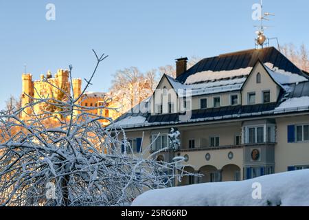 Bezauberndes Winterwunderland: Schneebedecktes europäisches Dorfhaus mit blauem Dach und mittelalterlichem Schloss im goldenen Sonnenlicht Stockfoto