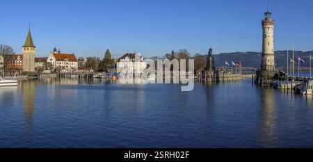 Panoramafoto des Hafens mit Mannturm, neuem Leuchtturm und bayerischem Löwen am Hafeneingang und mit Wasserspiegelung mit blauem Himmel im Stockfoto