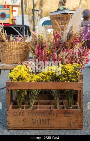 Eine rustikale Holzkiste voller lebendiger Blumen auf einem Markt im Freien, umgeben von Korbkörben und handgefertigten Arrangements. Montbrun-Bocage, Frankreich Stockfoto