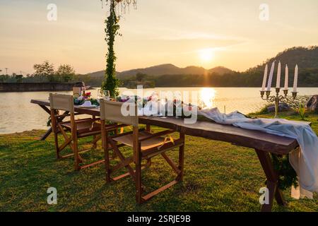 Rustikale und romantische elegante Outdoor-Dining-Einrichtung mit langem Holztisch, Stühlen, Geschirr, Besteck, Kerze und Flora Dekoration bei Sonnenuntergang Stockfoto