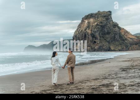 Asiatisches Paar macht einen Spaziergang am Strand mit Fußspuren und Lion Rock am Piha Beach in Auckland, Neuseeland Stockfoto