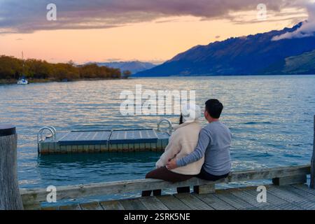 Rückansicht eines asiatischen Paares, das im Herbst auf dem Lake Wakatipu in Neuseeland sitzt und den Sonnenuntergang und den Berg genießt Stockfoto