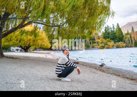 Schöne junge asiatische Frau, die sich unter Weiden am Seeufer hockt und im Herbst in Queenstown, Neuseeland, eine Herde Ente herstellt Stockfoto