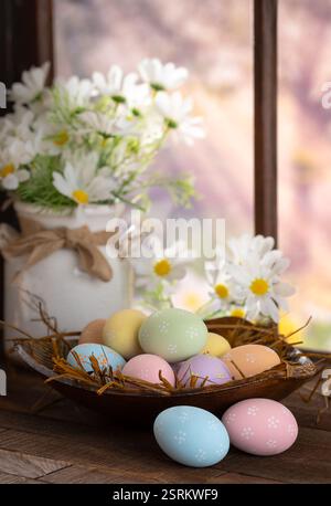 Ostereier in einer Schüssel mit weißen Gänseblümchen auf einem Holztisch neben dem Fenster mit farbenfrohem Frühlingshintergrund Stockfoto