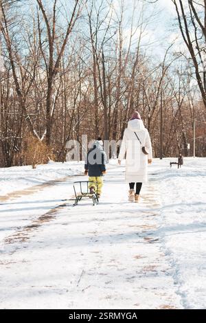 Unbekannte Mutter und Sohn mit Schlittenlaufen im Winterpark. Junge zieht Schlitten mit Mutter im schneebedeckten Park. Winteraktivität. Familienleben. Kleinkind mit Mutter Stockfoto