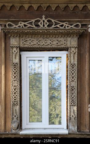 Ein weißes Fenster wird von aufwendigen Schnitzereien auf einem rustikalen Gebäude mit reflektiertem Grün eingerahmt. Stockfoto