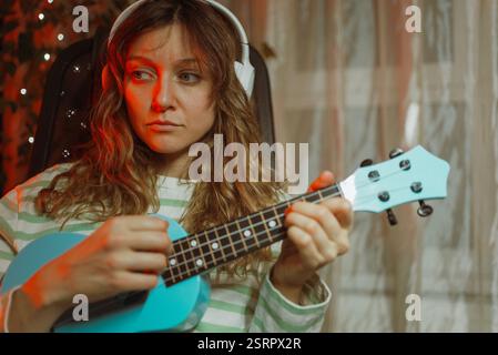 Eine junge Frau, die auf einem Stuhl sitzt, spielt eine hellblaue Ukulele, während sie weiße Kopfhörer trägt, mit roten Lichtern und einer Pflanze im Hintergrund Stockfoto