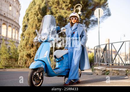 An einem sonnigen Tag in Rom, Italien, steht eine stilvolle Frau in blauem Jumpsuit und Mantel selbstbewusst neben ihrem Roller. Eine perfekte Darstellung italienischer Eleganz, Stadtleben und unabhängiges Reisen Stockfoto