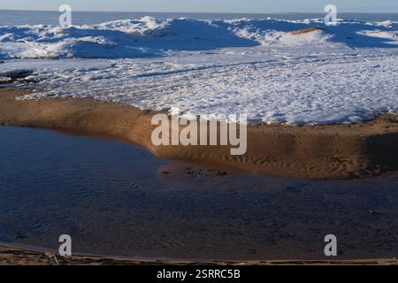 Tauender Bach, der in den Golf fließt, mit sichtbaren Sandküsten unter schmelzendem Schnee und Eis. Gefrorenes Meer im Hintergrund, wechselnde Jahreszeiten, instabiles Wetter Stockfoto