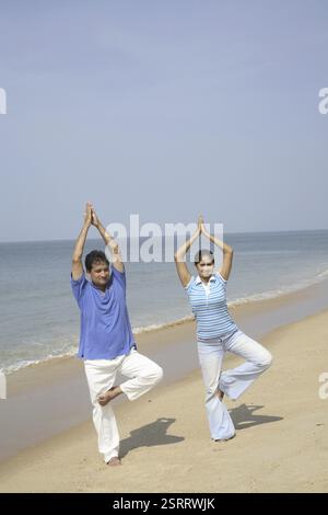 Südasiatische indische junge Mann und Frau, die Yoga machen, stehen auf einem gefalteten Bein und heben Gelenkhandhandflächen über der Küste, Shiroda, Dist. Sindhu Stockfoto