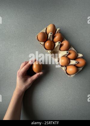 Braune Hühnereier in Papiertablett auf grauem Hintergrund mit einer Hand, die ein Ei hält Stockfoto