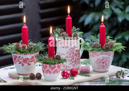 Adventsarrangements mit roten Kerzen, Nadelzweigen, Lärchenkegeln und Efeubeeren in Tassen Stockfoto