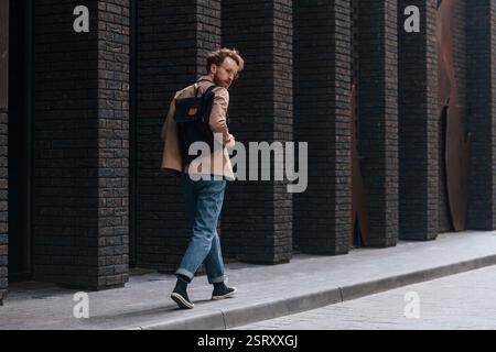 Stilvoller Mann mit Bart in khakifarbener Jacke und in Jeans ist draußen in der Nähe des Gebäudes. Stockfoto