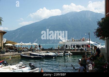 Einige Touristen gehen an einem sehr sonnigen Sommertag an einem kleinen Pier von Limone Sul Garda, Lago di Garda, Italien. Stockfoto