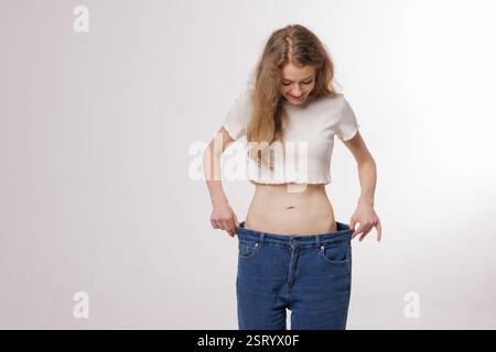 Form schlanke, dünne Taille, attraktive schlanke asiatische junge Frau, Hand zeigen ihre Gewichtsabnahme, tragen in großen, großen oder übergroßen Jeans, Übermaß verlieren durch Stockfoto