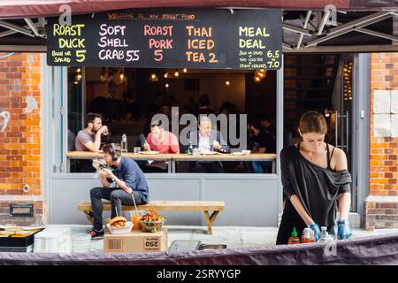 Leather Lane Market, London, bietet Entenbraten, Softshell-Krabben und thailändischen Eistee. Gäste speisen an Tischen. Stockfoto