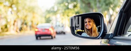 Glückliche schöne Frau mittleren Alters im Rückspiegel ihres neuen Autos. Horizontales Foto im Bannerformat mit Kopierraum. Stockfoto