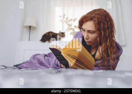 Eine Frau entspannt sich zu Hause und liegt mit einem Buch und einer Katze im Bett Stockfoto