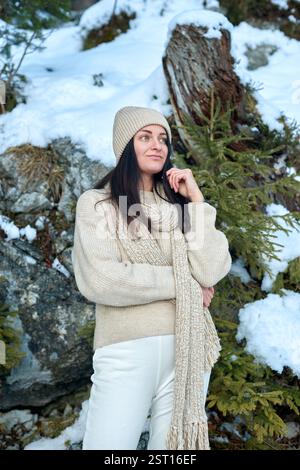 Junge Frau in cremefarbenem Strickpullover und Beanie, die in einer ruhigen, verschneiten Berglandschaft mit felsigen Felsvorsprüngen und immergrünen Bäumen steht Stockfoto
