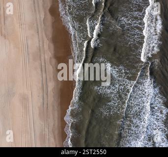Luftaufnahme von Wellen, die mit Reifenspuren im Sand brechen Stockfoto