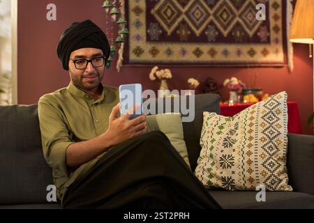 Mann im Turban sitzt auf der Couch und lächelt, während er ein Smartphone im gemütlichen Wohnzimmer mit gemusterten Kissen und Wanddekor benutzt Stockfoto