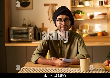Porträt eines jungen Mannes mit einer Brille, der Turban trägt, am Küchentisch sitzt, mit einem Tablet in den Händen warm lächelt, direkt in die Kamera blickt und eine freundliche Atmosphäre schafft Stockfoto