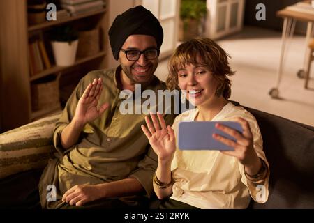 Zwei Freunde sitzen auf der Couch, während sie mit dem Smartphone winken und Videoanrufe im Wohnzimmer tätigen. Ein Freund trägt Turban, der andere hält das Telefon Stockfoto