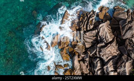 Granitfelsen mit Meereswellen, die gegen sie plätschern, umgeben von lebendigem blauem Wasser. Seychellen, La Digue. Stockfoto