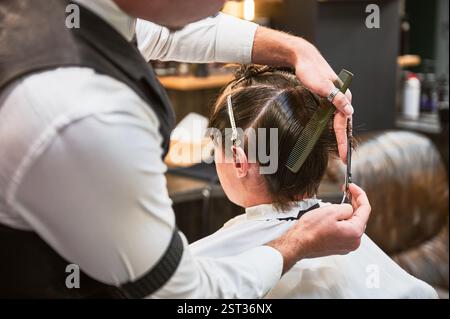 Rückansicht eines Friseurs im schicken Outfit, der nasse Haare schneidet. Junger Kunde bekommt einen stilvollen Haarschnitt. Frisches Styling vom professionellen Friseur. Stockfoto