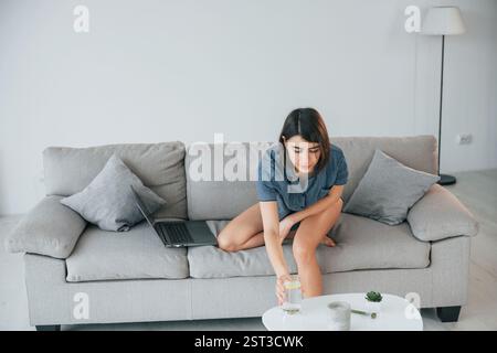 Auf dem Sofa sitzen und Glas mit Wasser nehmen. Die junge Frau ist zu Hause. Vorstellung von Schönheit. Stockfoto
