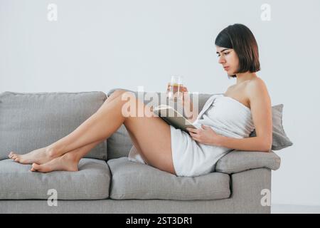 Legen Sie sich auf das Sofa und lesen Sie das Buch, während Sie das Glas mit Wasser halten. Die junge Frau ist zu Hause. Vorstellung von Schönheit. Stockfoto
