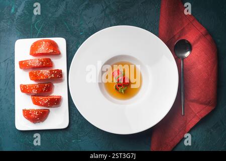 Konsomme oder Brühe mit Lachsbrötchen serviert auf einem weißen Teller mit einem roten Sesambrötchen auf dunkelgrünem Hintergrund mit Serviette und Löffel. Hochwertige Pho Stockfoto