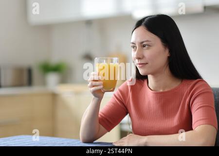 Asiatische Frau, die Orangensaft hält und zu Hause in der Küche nachdenkt Stockfoto