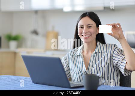 Glückliche asiatische Frau, die einen Laptop benutzt und in der Küche zu Hause eine leere Karte zeigt Stockfoto