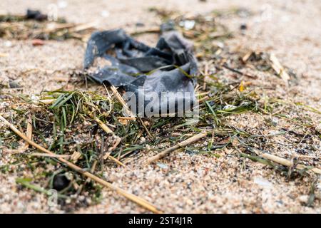 Ein Stück alten Stoffes, das auf dem Meer entsorgt wird, liegt am Sandstrand zwischen Algen und Muscheln, was die Abfälle der Textilindustrie veranschaulicht, die den Ozean verschmutzen Stockfoto
